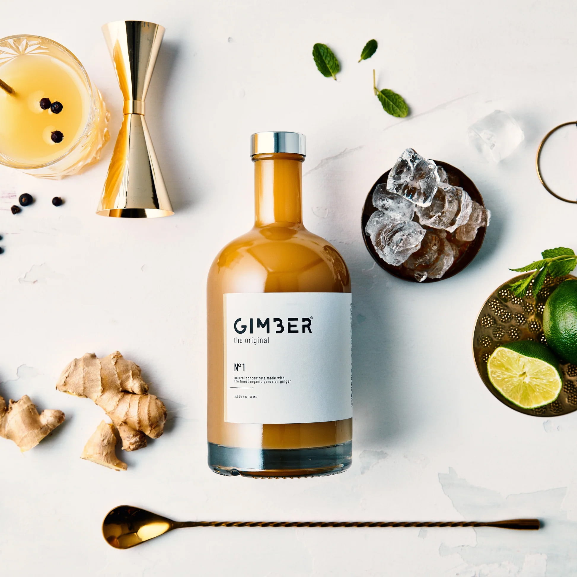 Une bouteille de concentré de gingembre GIMBER est entourée d'un verre à cocktail, d'un jigger, de racines de gingembre, de glaçons, de citron vert, de feuilles de menthe, d'une cuillère dorée et de grains de poivre noir sur une surface blanche lumineuse de type cantine.