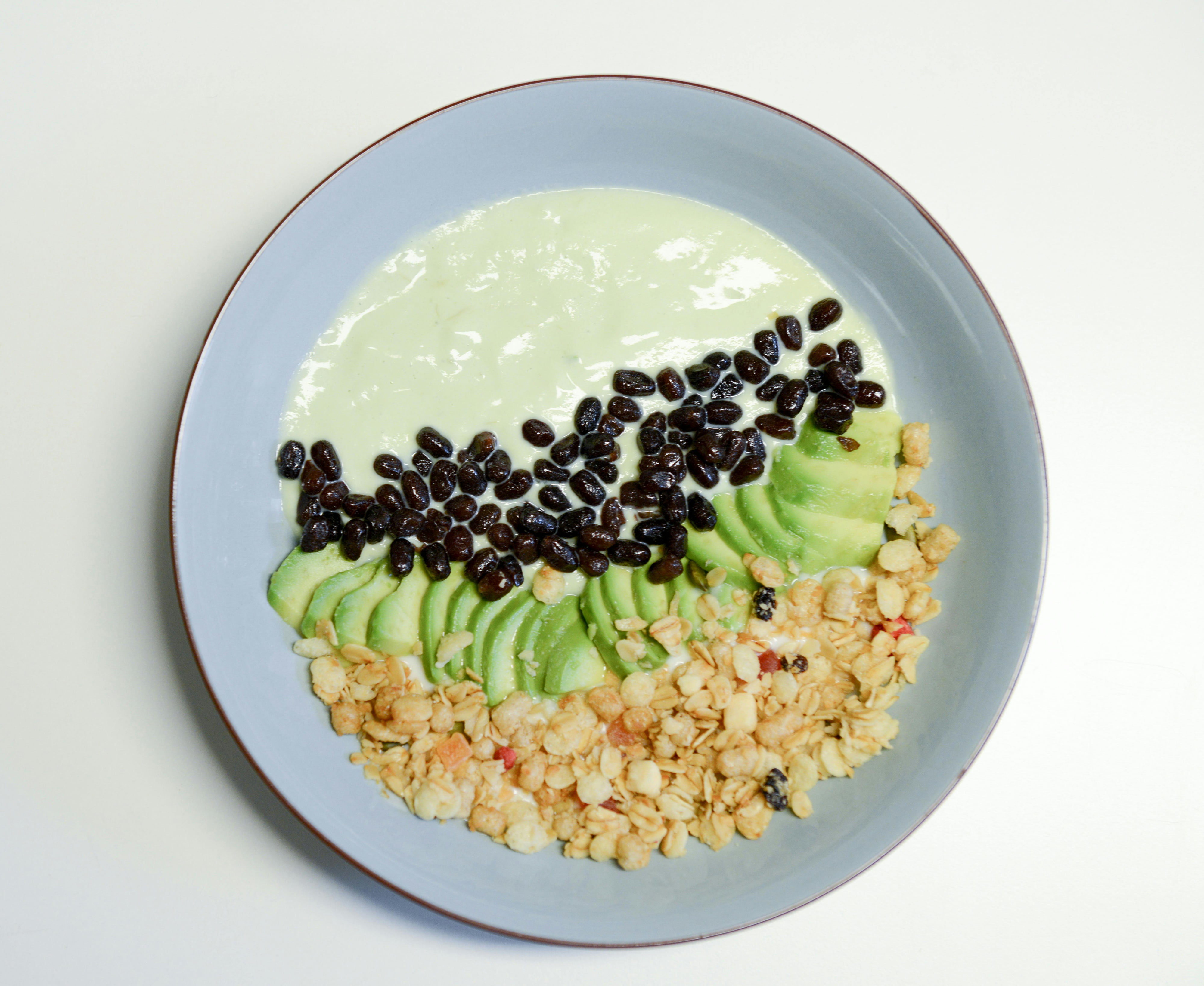 Un bol de yaourt garni de tranches d'avocat, de haricots noirs et de granulés - disposés en trois sections distinctes - apporte une touche de rebranding à un style cantine lumineux sur un fond blanc épuré.
