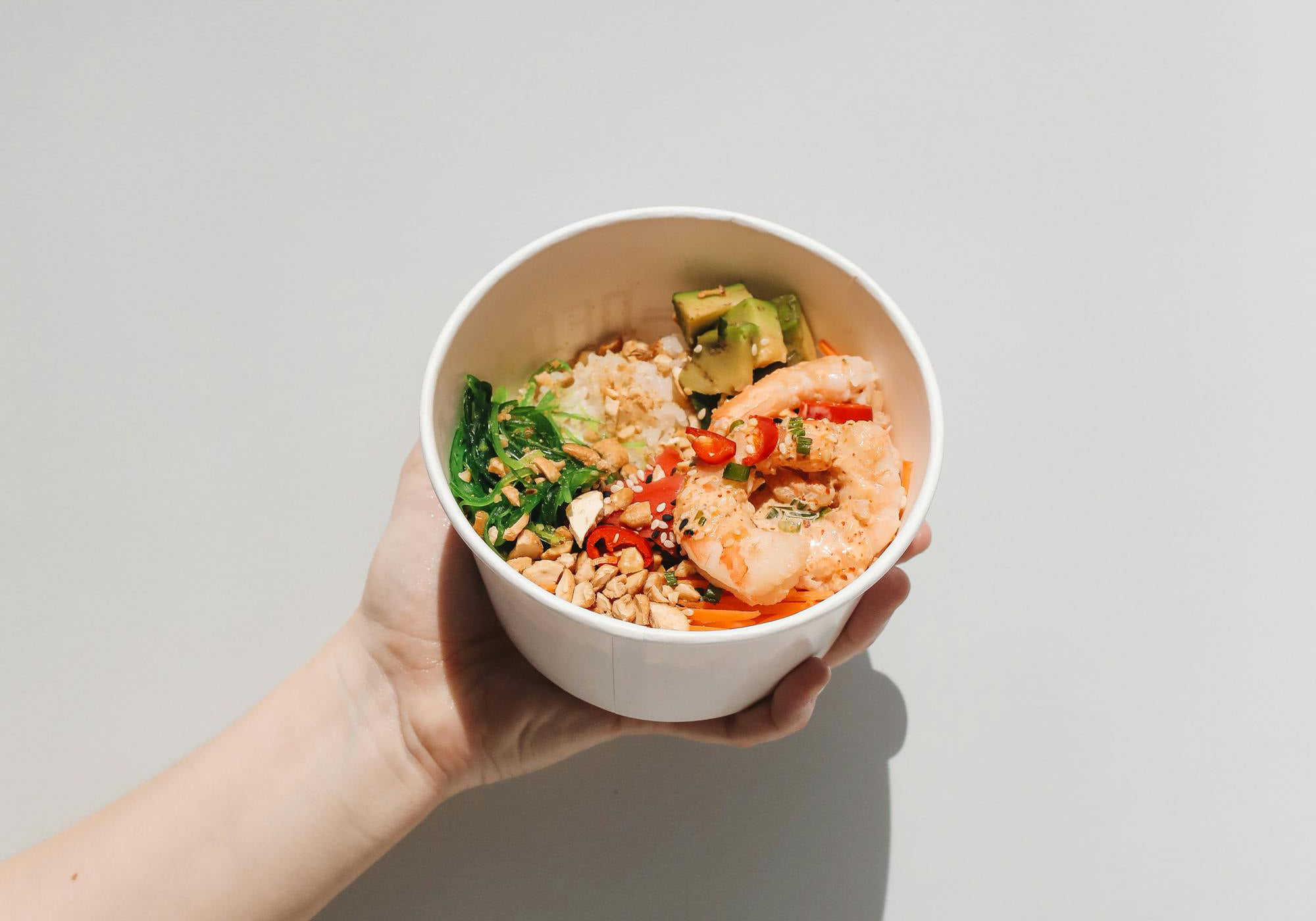 Une main tient un bol blanc issu du rebranding de Bright Canteen, rempli de crevettes, de riz, de légumes hachés, de cacahuètes, d'algues vertes et de graines de sésame sur un fond blanc uni.