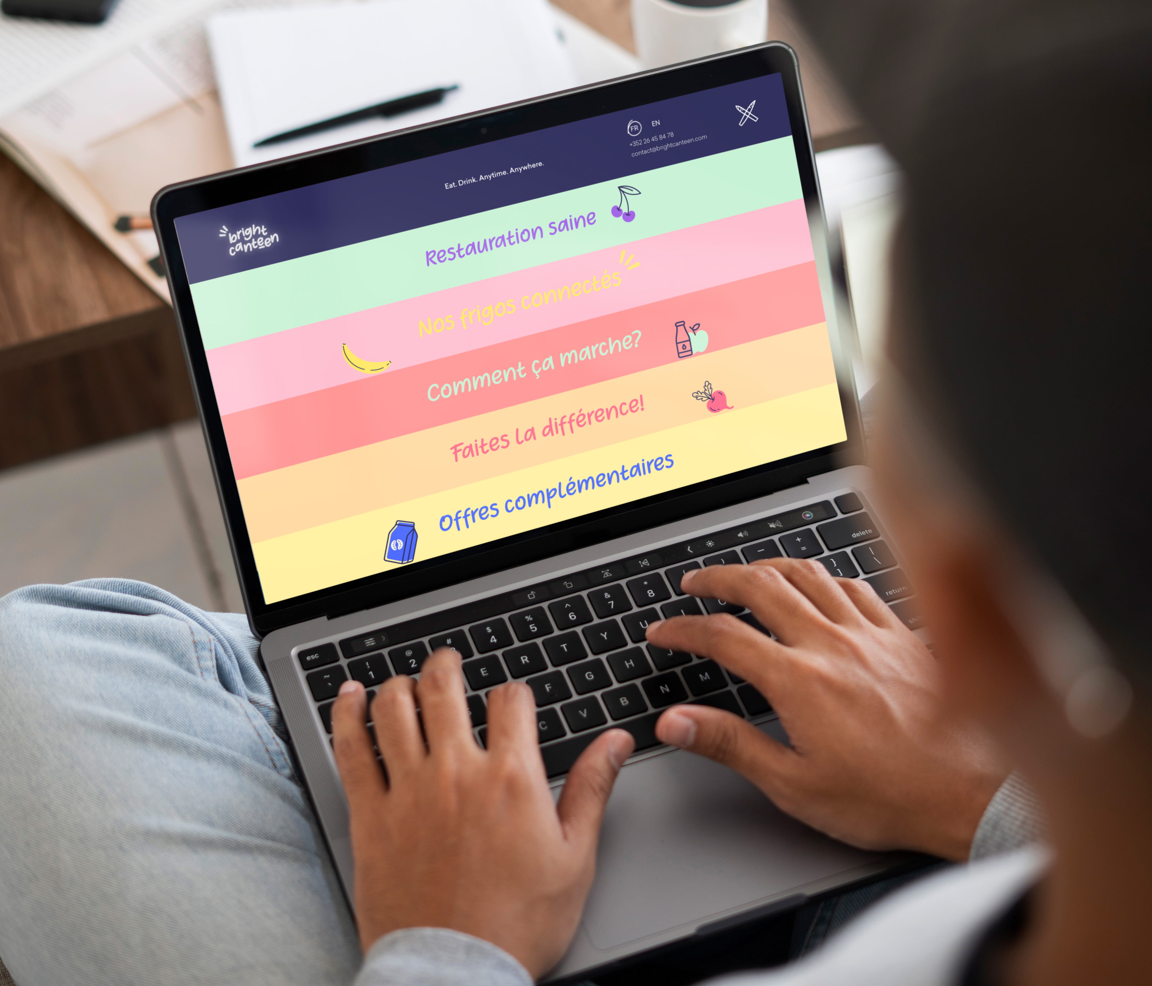 Une personne tape sur un ordinateur portable qui affiche la nouvelle image de Takaneo : un site web lumineux, en français, avec des sections sur l'alimentation saine, des questions courantes et des offres supplémentaires, avec des icônes ludiques et des arrière-plans pastel inspirés des cantines.
