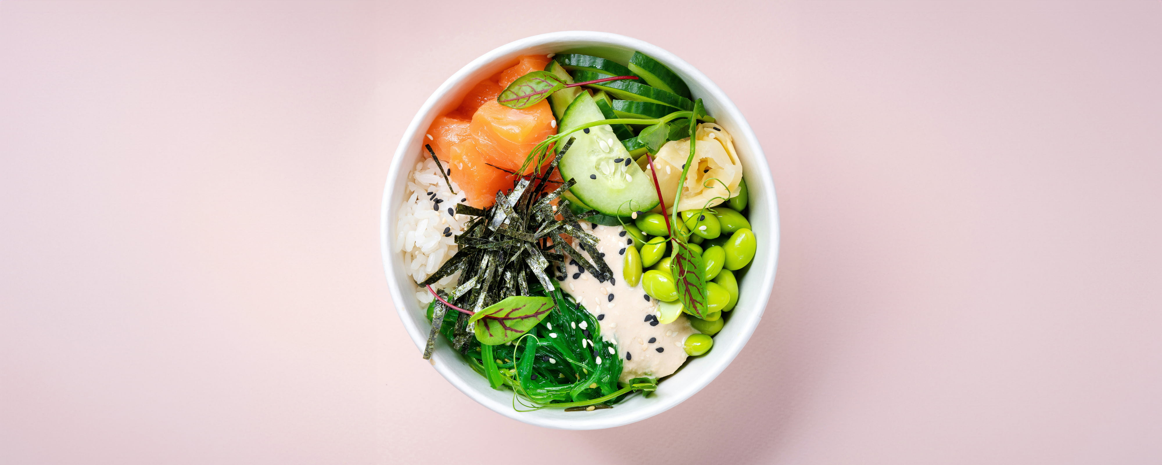Un classique revisité : ce bol poké lumineux contient du riz blanc, des cubes de saumon, des algues, du concombre, des edamames, des légumes verts, de la sauce et des graines de sésame dans un bol blanc sur fond rose pâle - un choix parfait pour votre cantine ou votre repas d'inspiration takaneo.