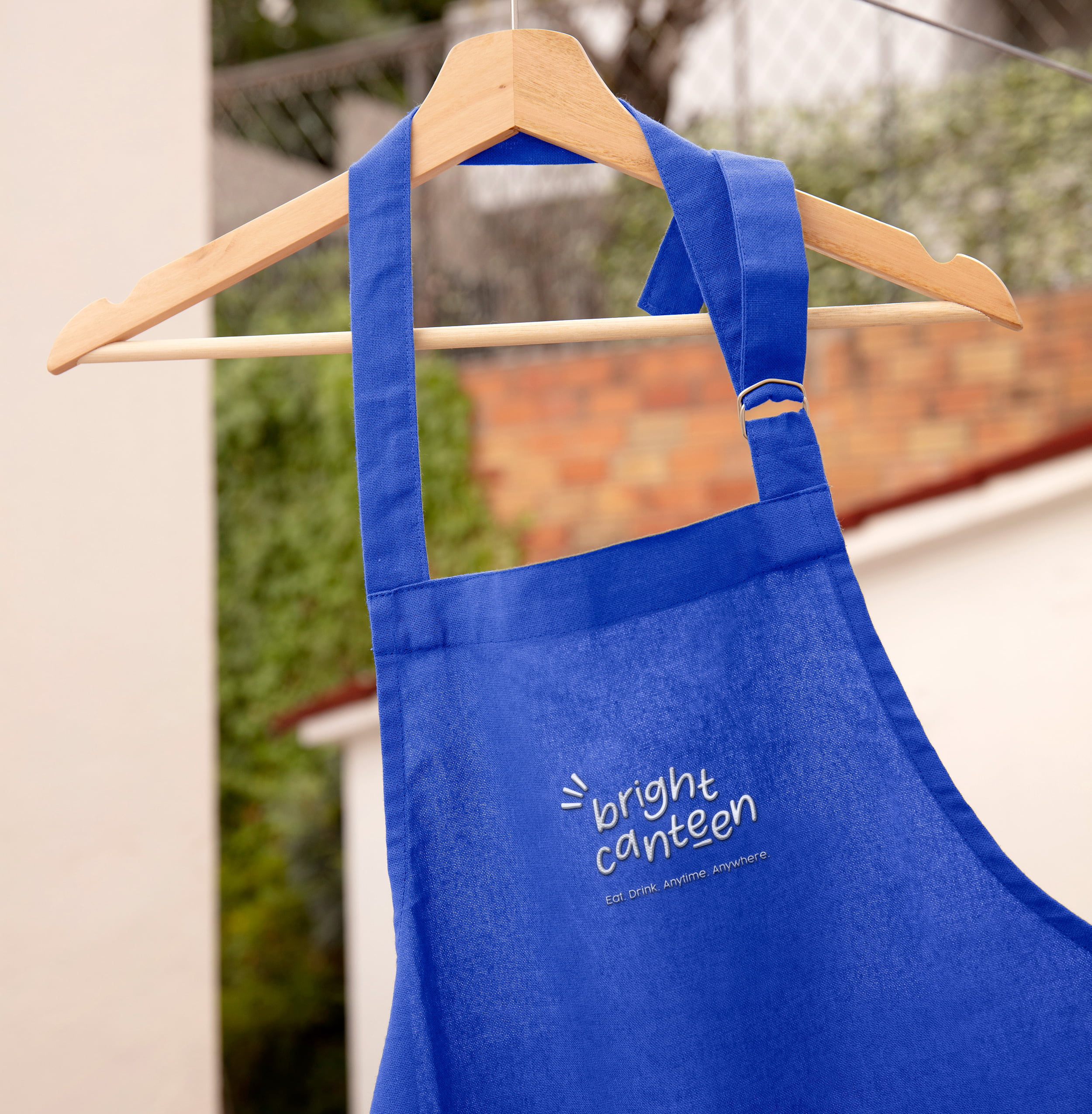Un tablier bleu royal portant l'inscription bright canteen : eat. drink. anywhere. anytime - un clin d'œil au rebranding de Bright Canteen - est suspendu à un cintre en bois à l'extérieur, avec un mur de briques floues et de la verdure en arrière-plan.