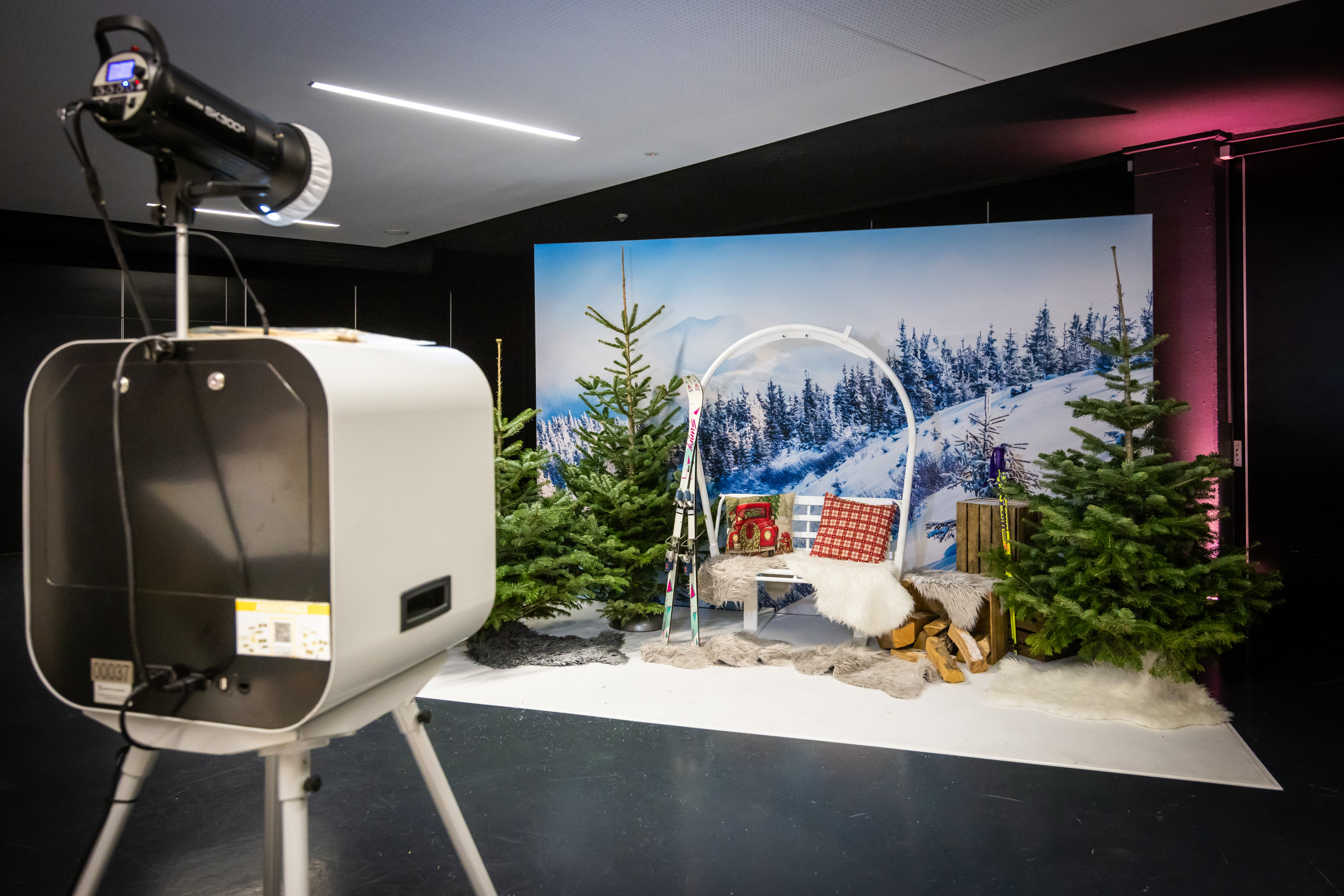 Un photomaton est installé sur une toile de fond hivernale avec des arbres enneigés, des sapins décorés, de la fausse neige, une chaise blanche et des accessoires d'hiver. Un grand éclairage de studio et un appareil photo sont placés au premier plan.