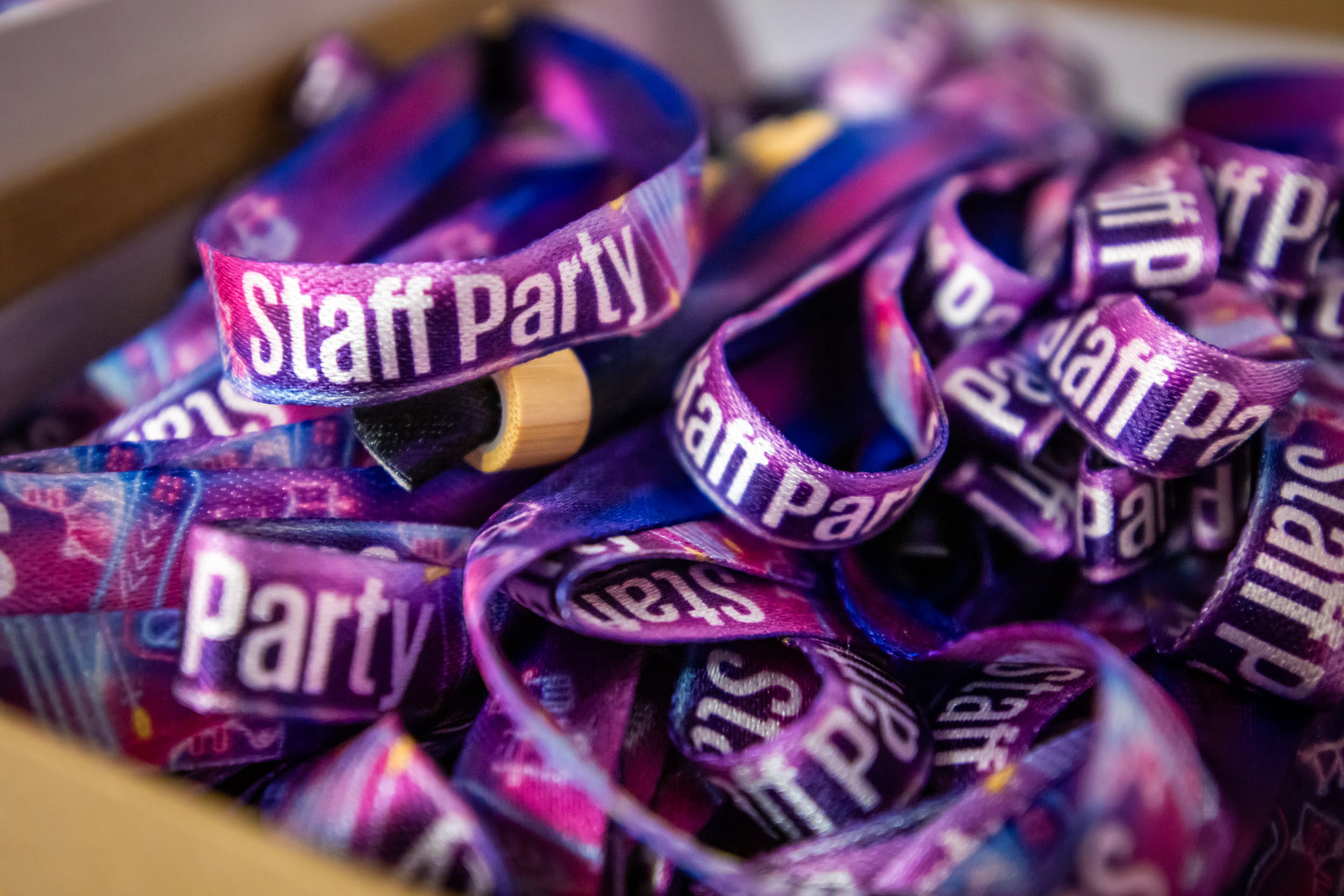 Gros plan sur plusieurs bracelets en tissu coloré sur lesquels sont imprimés en blanc les mots Staff Party (fête du personnel). Les bracelets sont violets avec des touches de bleu et de rose et sont empilés dans une boîte.