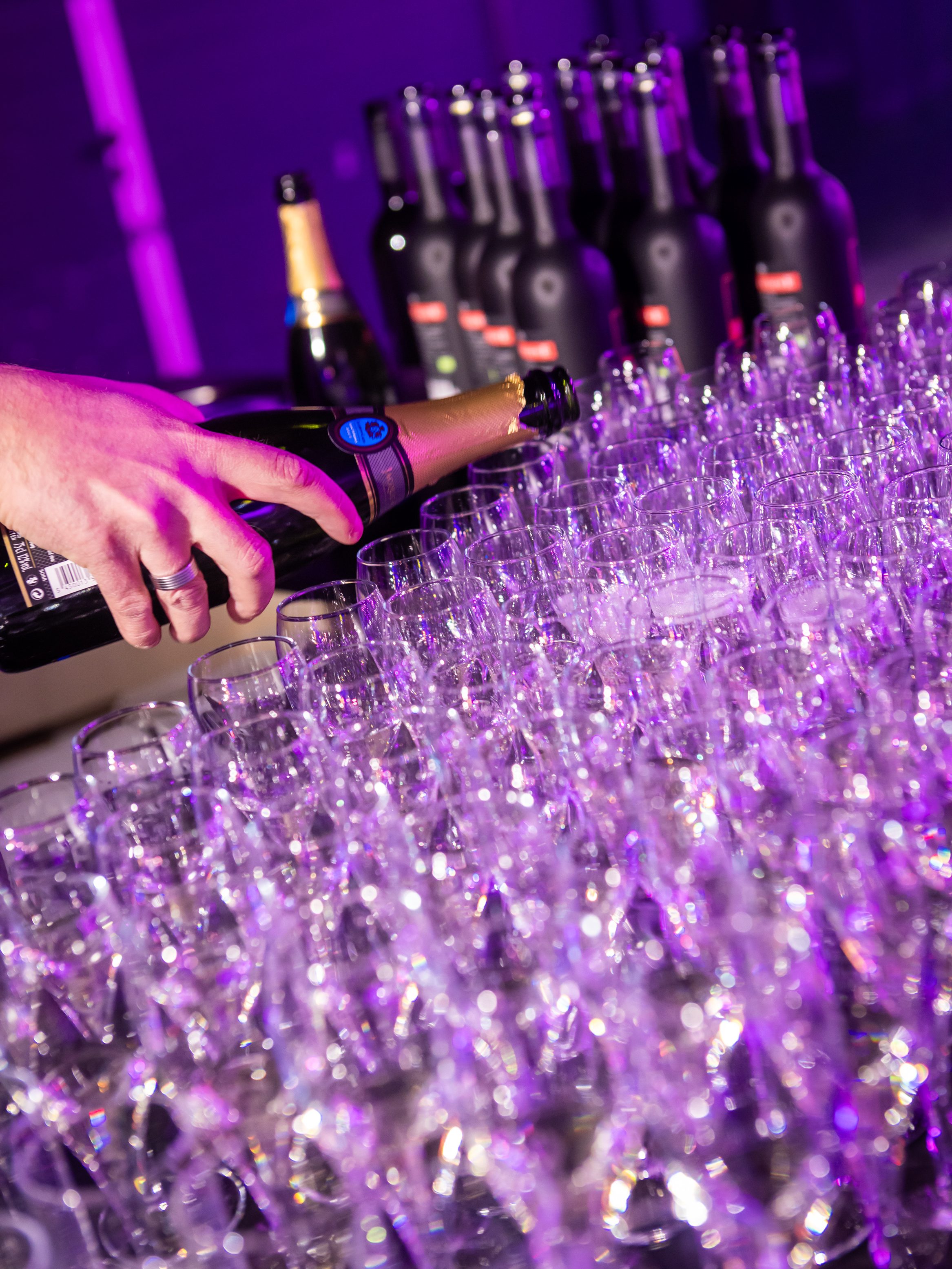 Une personne verse du champagne dans une rangée de verres vides sur une table, avec plusieurs bouteilles de vin et de champagne en arrière-plan, le tout éclairé par une lumière violette - parfait pour capturer des photos et vidéos d'entreprise lors d'un évènement.