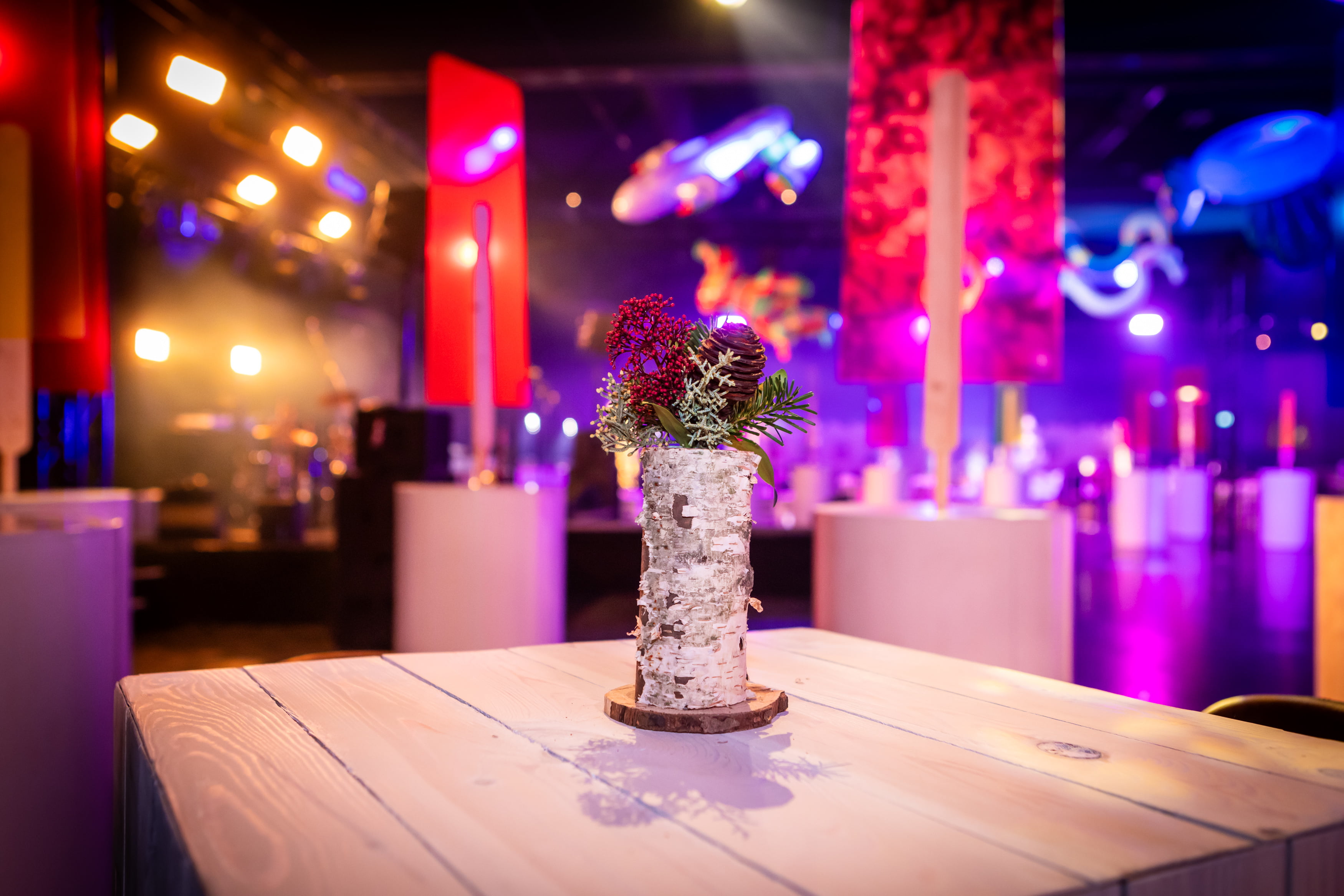 Un vase en bouleau avec du feuillage rouge et vert est posé sur une table en bois clair dans un espace événementiel coloré et doucement éclairé, avec des lumières et des décorations floues en arrière-plan.
