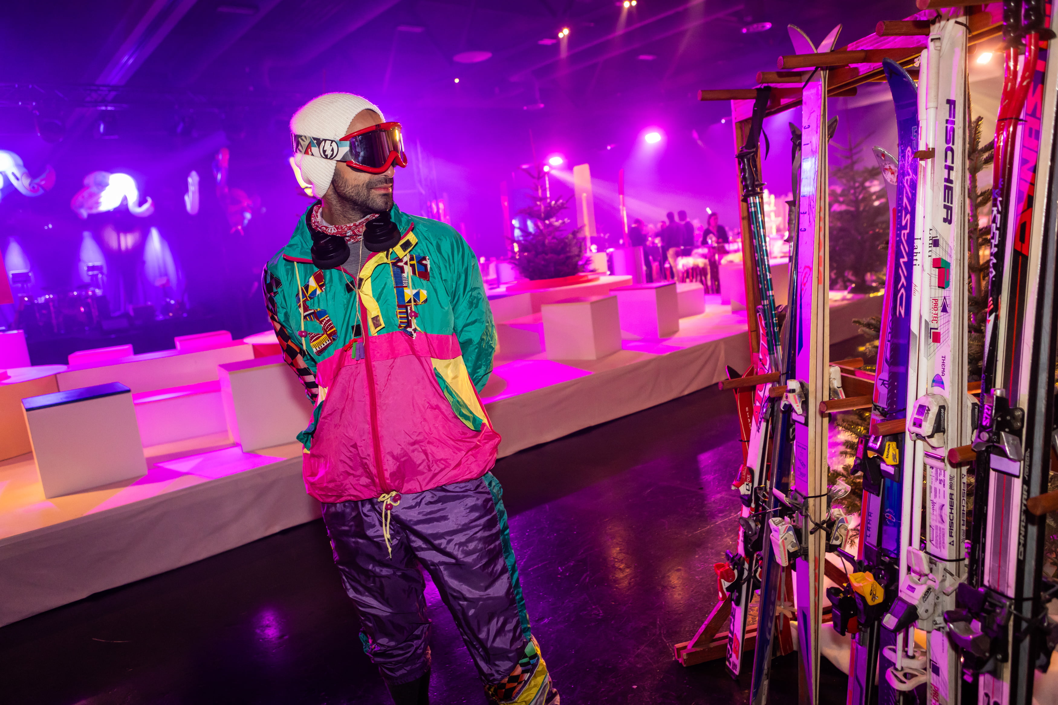 Un homme portant une tenue de ski rétro colorée et des lunettes de ski se tient à l'intérieur près d'un rack de skis vintage, avec un éclairage violet vibrant et des sièges de salon décorés à l'arrière-plan.