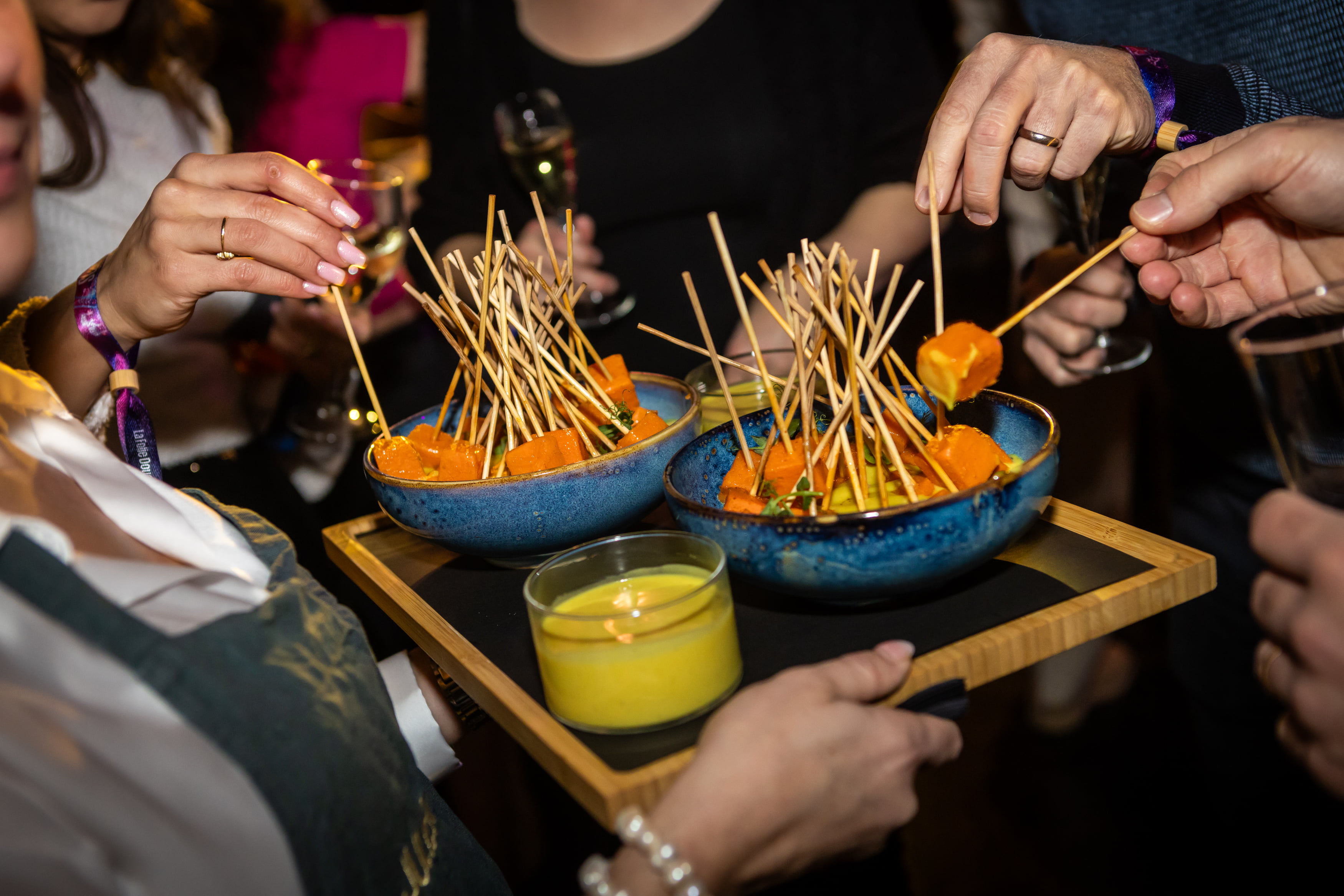 Plusieurs personnes prennent des amuse-gueules en brochettes dans des bols bleus sur un plateau lors d'une réunion sociale. Une bougie jaune se trouve également sur le plateau et des personnes tiennent des boissons à l'arrière-plan. Le cadre semble animé et festif.