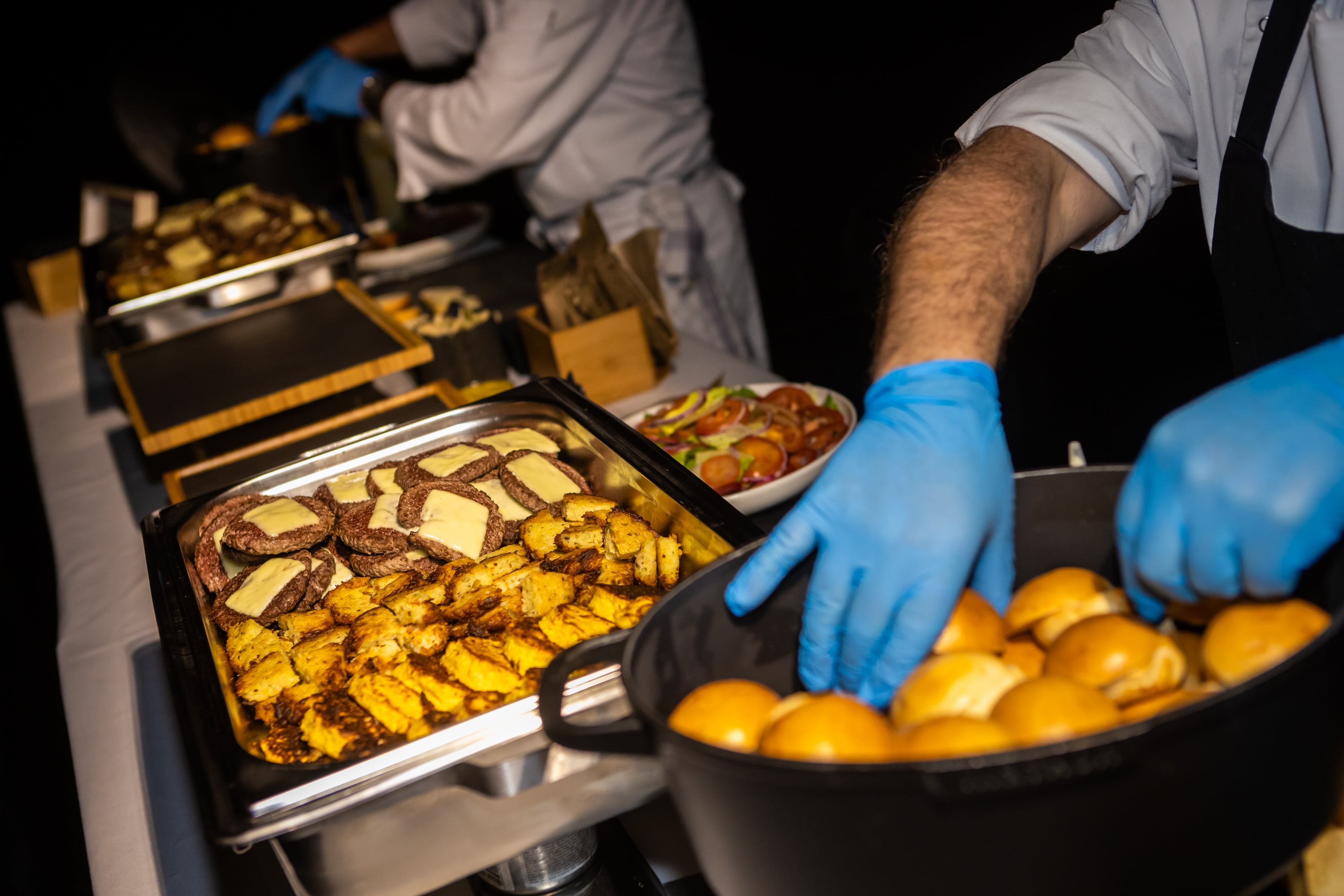 Un chef portant des gants bleus dispose des petits pains à côté d'un plateau de pommes de terre cuites et de viande grillée avec du fromage fondu dans une station de restauration de type buffet.