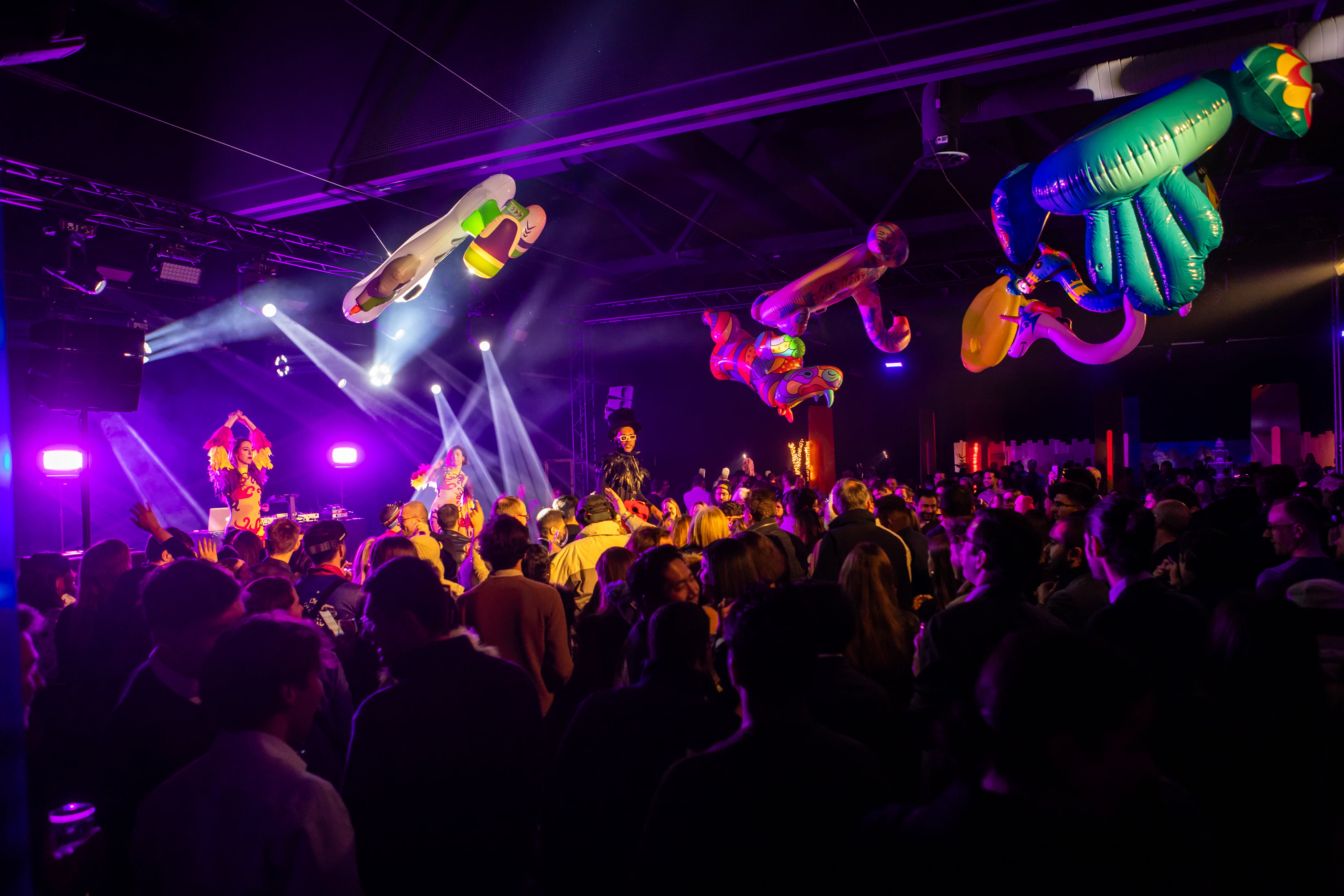 Une foule de personnes lors d'une fête ou d'un concert en intérieur, avec des lumières de scène colorées, des artistes en costumes éclatants et de grandes décorations gonflables suspendues au plafond.