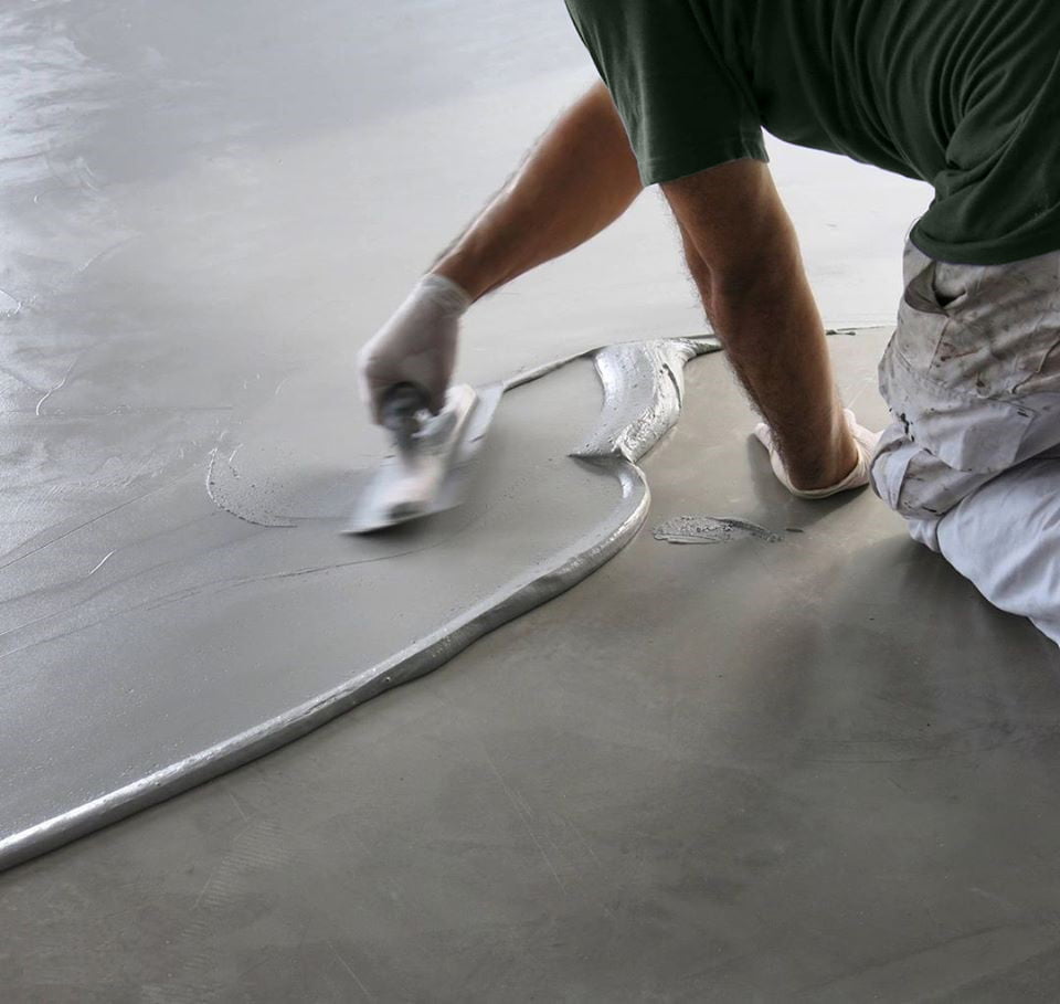 Une personne portant des gants s'agenouille sur le sol et utilise une truelle pour étaler et lisser le béton Trendy Stone humide sur une surface plane.