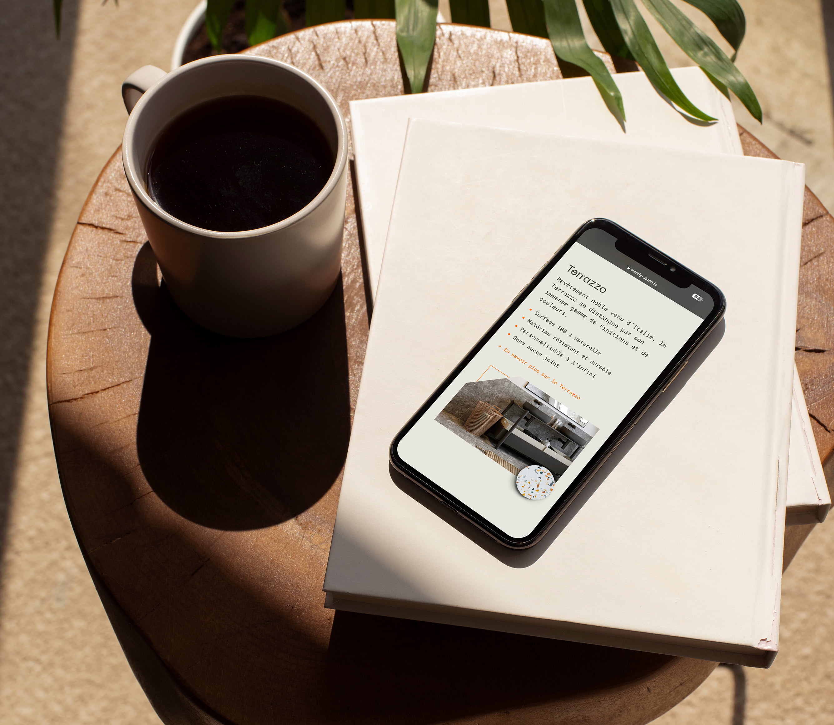 Un smartphone affichant une liste de propriétés Trendy Stone avec un logo de rebranding repose sur deux livres blancs empilés à côté d'une tasse de café noir, le tout sur une table ronde en bois, tandis que la lumière du soleil et les ombres des plantes à feuilles créent un réseau de motifs.