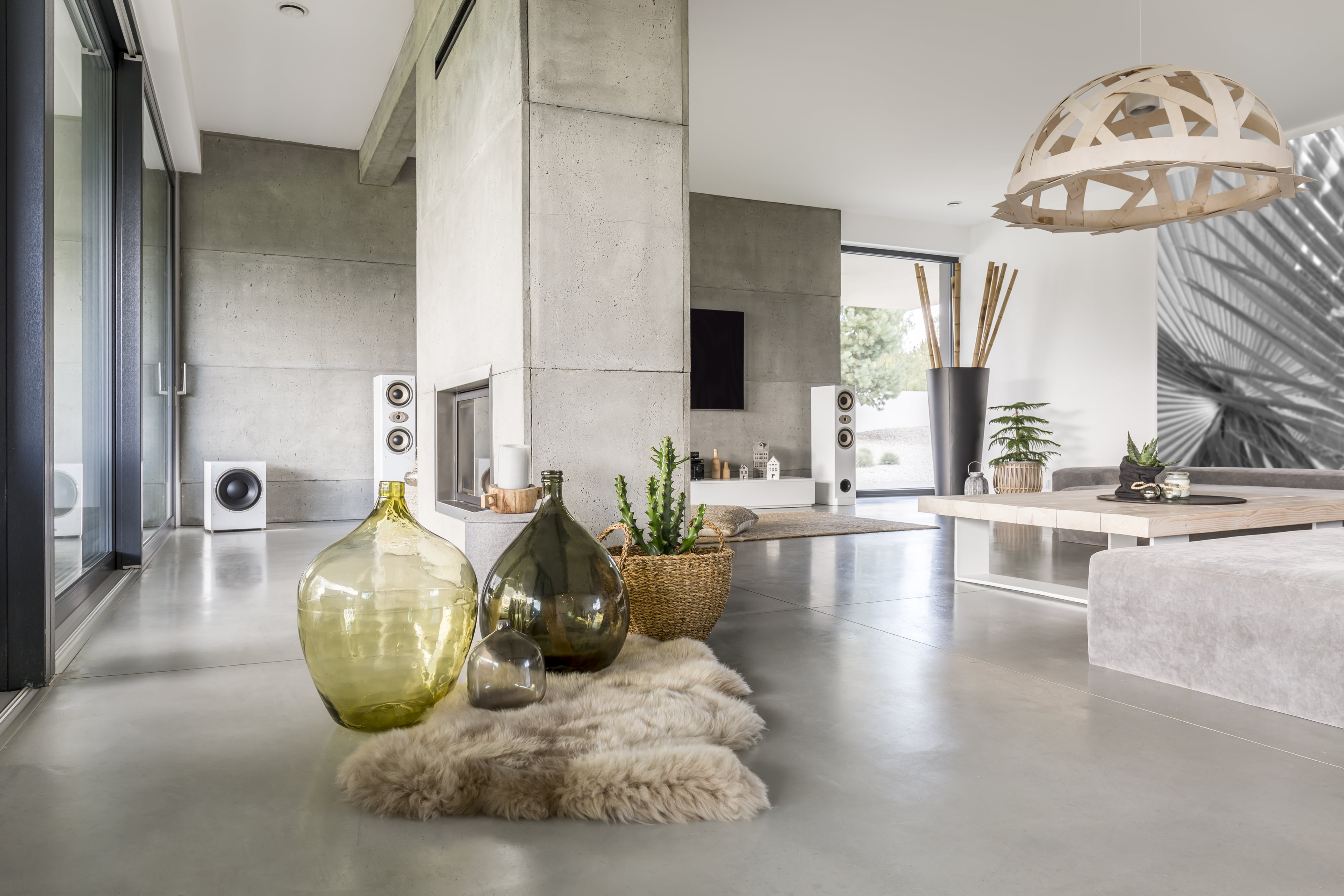 Salon moderne avec des murs et des sols en béton, de grandes fenêtres, des meubles minimalistes et une cheminée centrale. La décoration Stone tendance comprend des vases en verre décoratifs sur un tapis en fourrure, des plantes d'intérieur et une lampe suspendue en bois au-dessus de la table à manger.