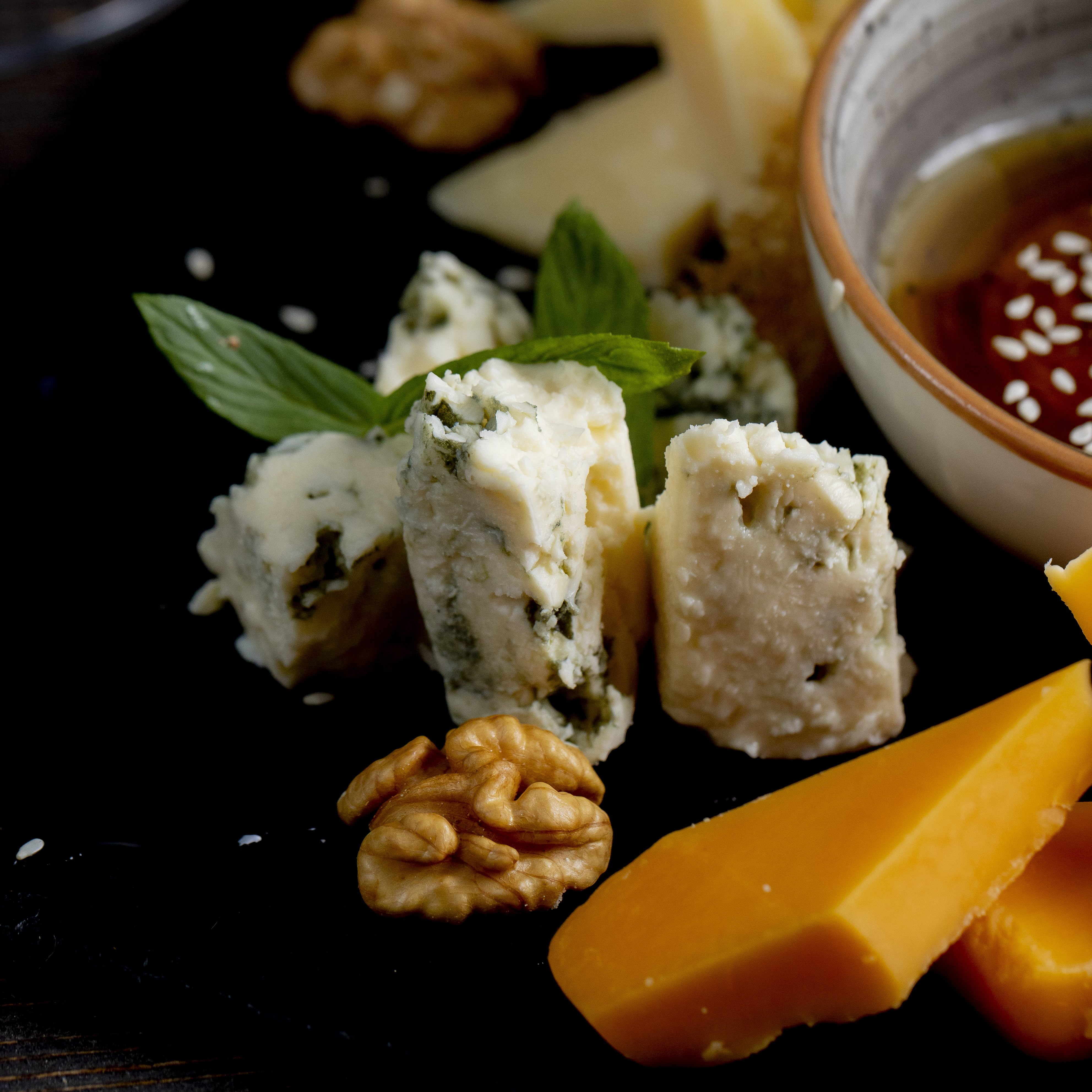 Gros plan sur un assortiment de fromages, de noix et d'une sauce dans un bol, garni de feuilles de menthe fraîche, disposés sur un plateau noir.