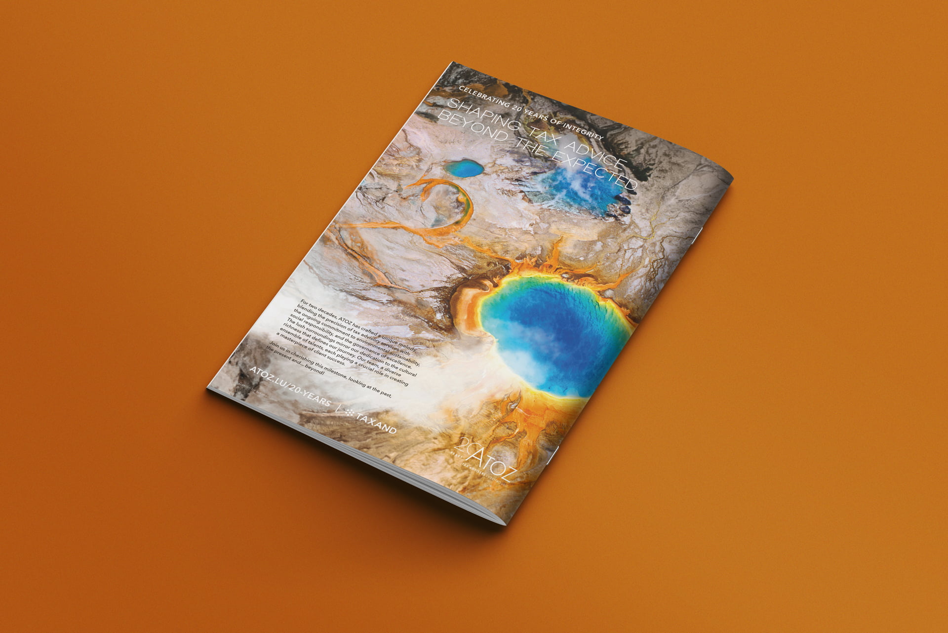 Un magazine célébrant les 20 ans d'Atoz repose sur une surface orange, sa couverture présentant une photo aérienne d'une source d'eau chaude et de formations minérales. Le texte blanc présente des titres d'articles sur des sujets liés au développement durable.