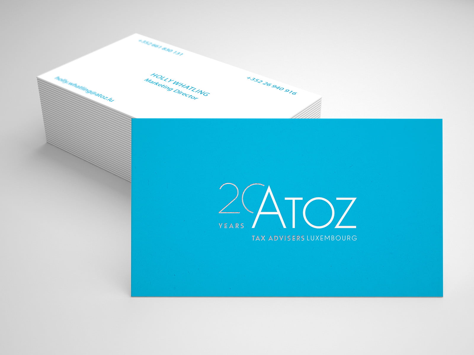 Une pile de cartes de visite avec un recto bleu vif indiquant 20 Years Atoz Tax Advisers Luxembourg celebrates Atoz 20 ans, et un verso blanc affichant les coordonnées durables de Holly Whittaker, Managing Director, en texte bleu.