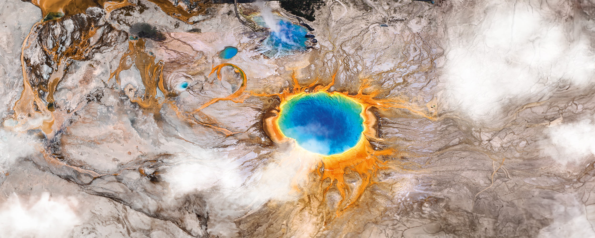 Vue aérienne du Grand Prismatic Spring dans le parc national de Yellowstone, capturée par Atoz 20 ans, montrant des anneaux vibrants de bleu, de vert, de jaune et d'orange au milieu d'un terrain rocailleux et de taches de nuages dans ce paysage durable.