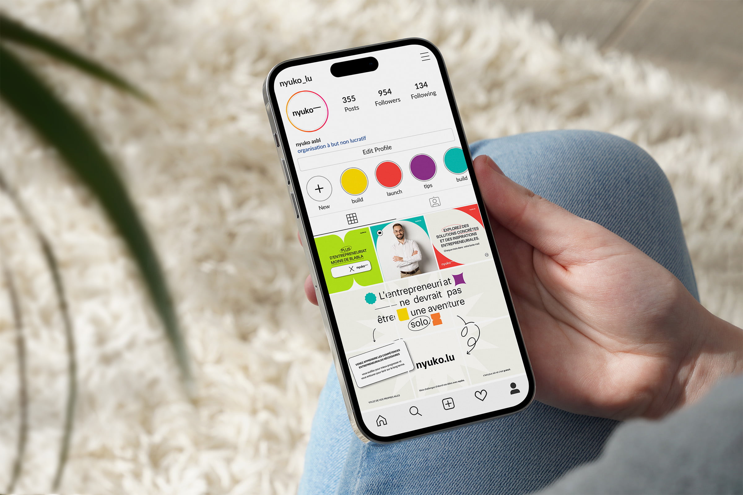 Une personne tient un smartphone affichant un profil Instagram avec des posts colorés, y compris des graphiques, des citations et une photo d'une personne. L'arrière-plan montre un tapis beige doux et un jean bleu.