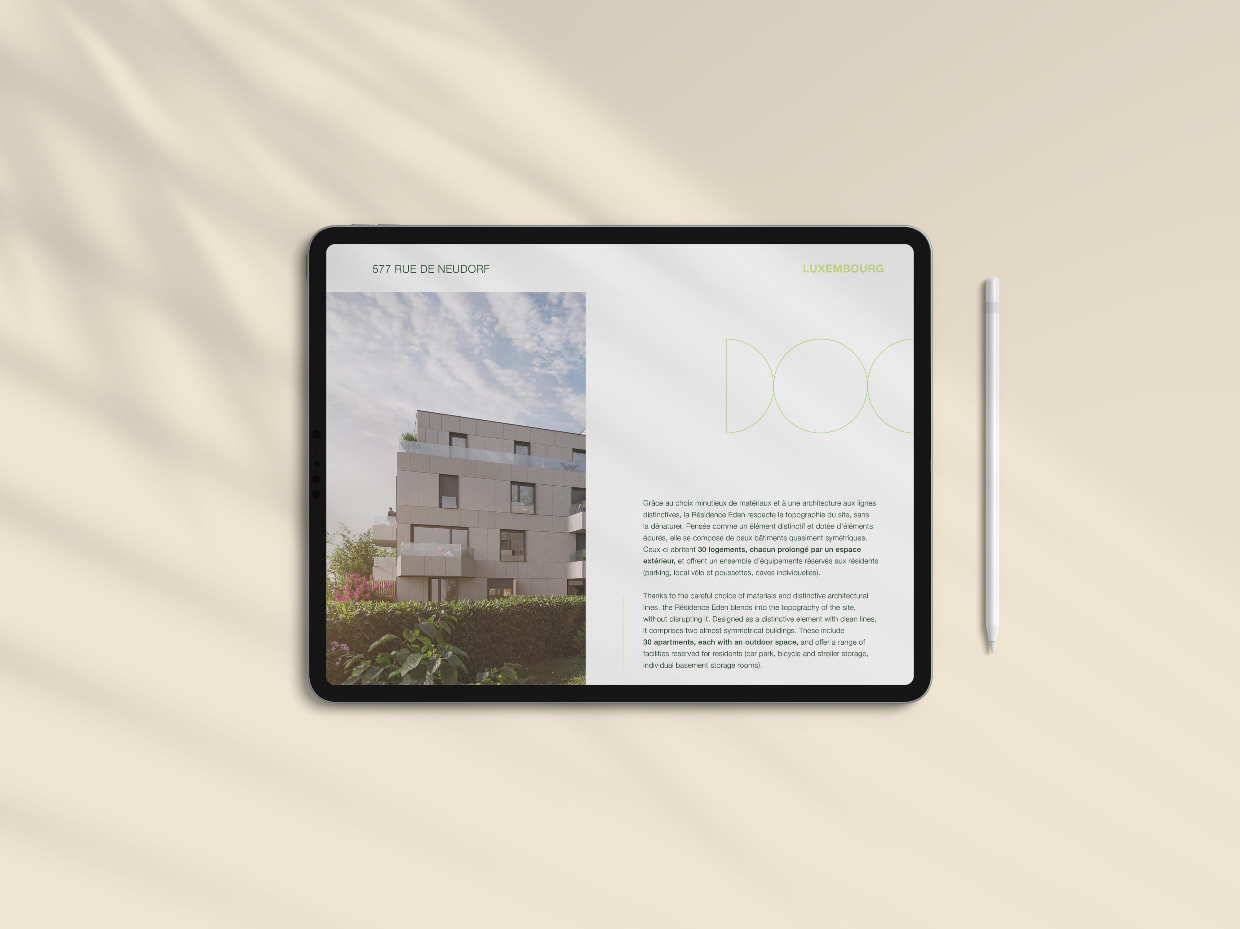Une tablette affichant une présentation d'architecture moderne avec une photo de bâtiment à gauche et un texte descriptif à droite, posée sur une surface beige à côté d'un stylet. Des ombres de feuilles sont visibles sur la surface.