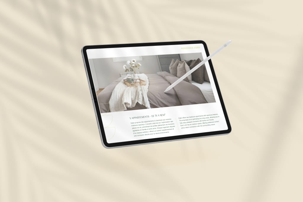 Une tablette avec un stylet affiche une page web de décoration d'intérieur présentant un lit soigneusement fait avec une literie beige et des oreillers décoratifs. L'arrière-plan est un beige doux et flou avec des ombres de feuilles.