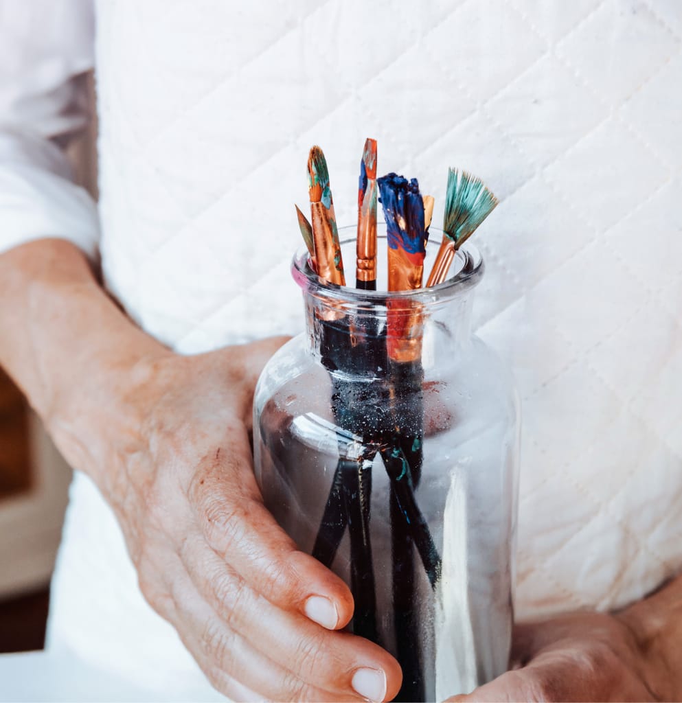 Une personne vêtue d'une chemise blanche matelassée tient un pot en verre transparent rempli de pinceaux colorés recouverts de peinture. Les mains et le pot sont mis au point, tandis que l'arrière-plan est flou.