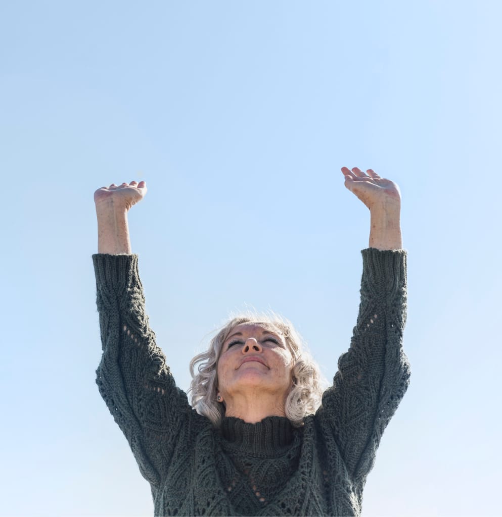 Une femme âgée aux cheveux gris, vêtue d'un pull-over vert, se tient à l'extérieur, souriante et levant les deux bras joyeusement vers un ciel bleu clair.