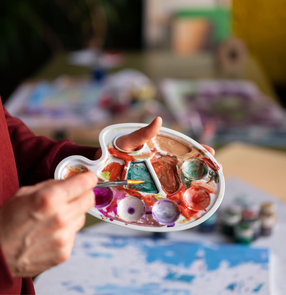 Une personne tient dans une main une palette de peinture remplie de différentes couleurs et utilise un pinceau pour mélanger la peinture. Des fournitures artistiques et des œuvres d'art colorées sont visibles sur une table à l'arrière-plan flou.
