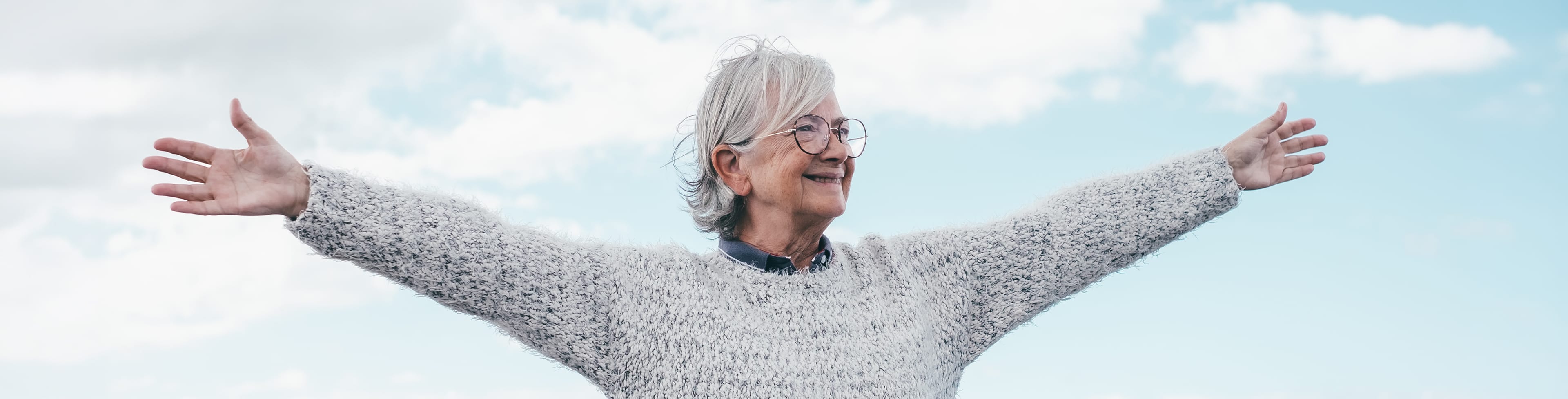 Une personne âgée aux cheveux gris et aux lunettes se tient en plein air dans un ciel bleu parsemé de nuages, souriante, les bras tendus, vêtue d'un pull-over gris confortable.