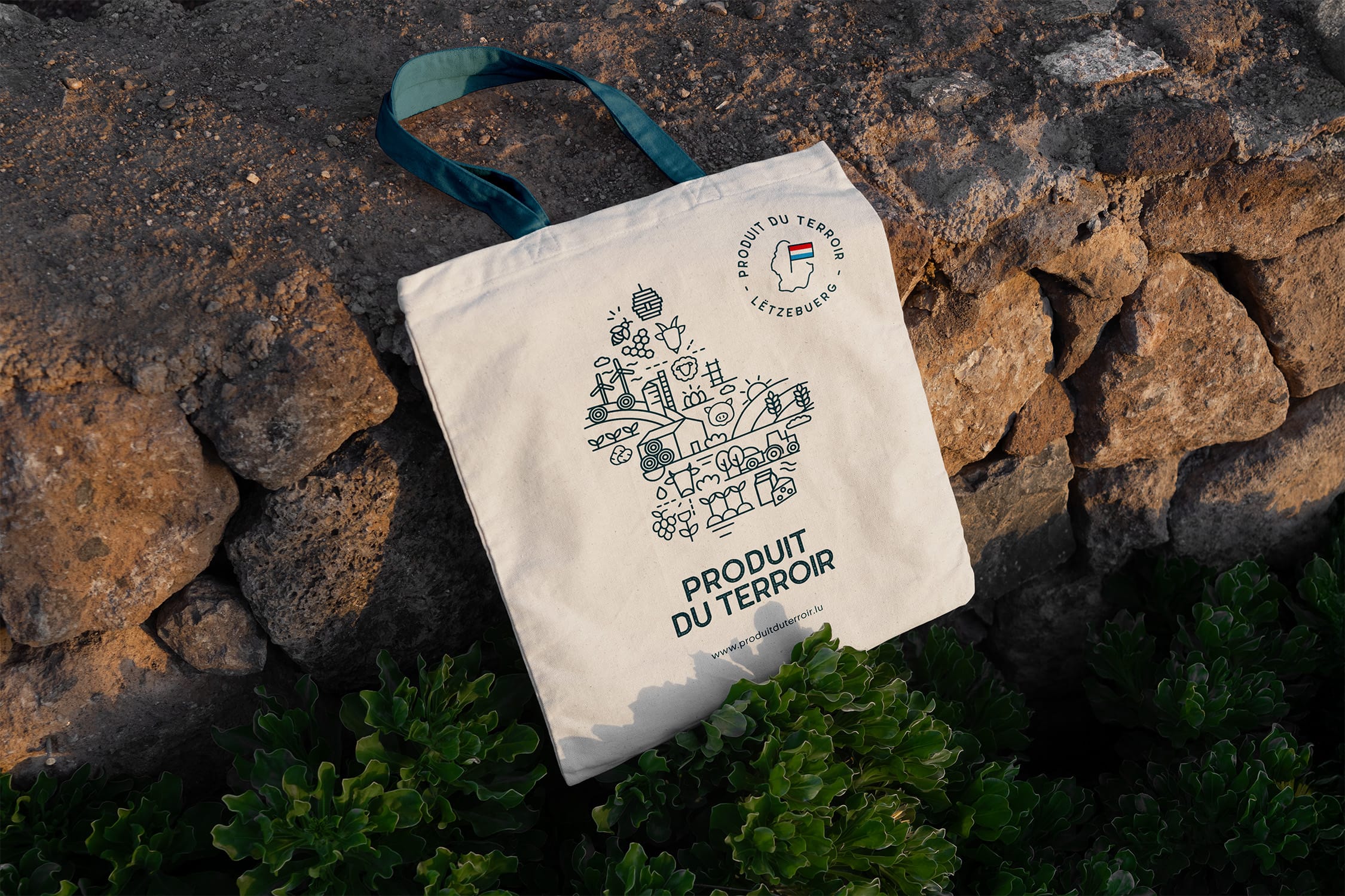 Un sac fourre-tout blanc avec des poignées vertes est posé contre un mur de pierre, affichant des symboles illustrés de l'alimentation et de l'agriculture et le texte PRODUIT DU TERROIR avec un logo circulaire au-dessus. Des plantes vertes poussent sous le sac.