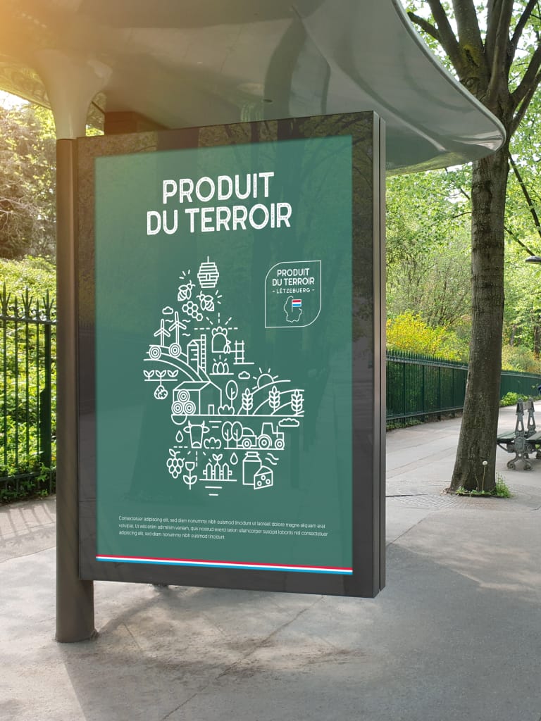 Sur l'affiche d'un arrêt de bus, on peut lire PRODUIT DU TERROIR au-dessus d'un arbre stylisé composé de dessins au trait blanc représentant des aliments, des animaux et des symboles agricoles, avec de la verdure et un sentier à l'arrière-plan.