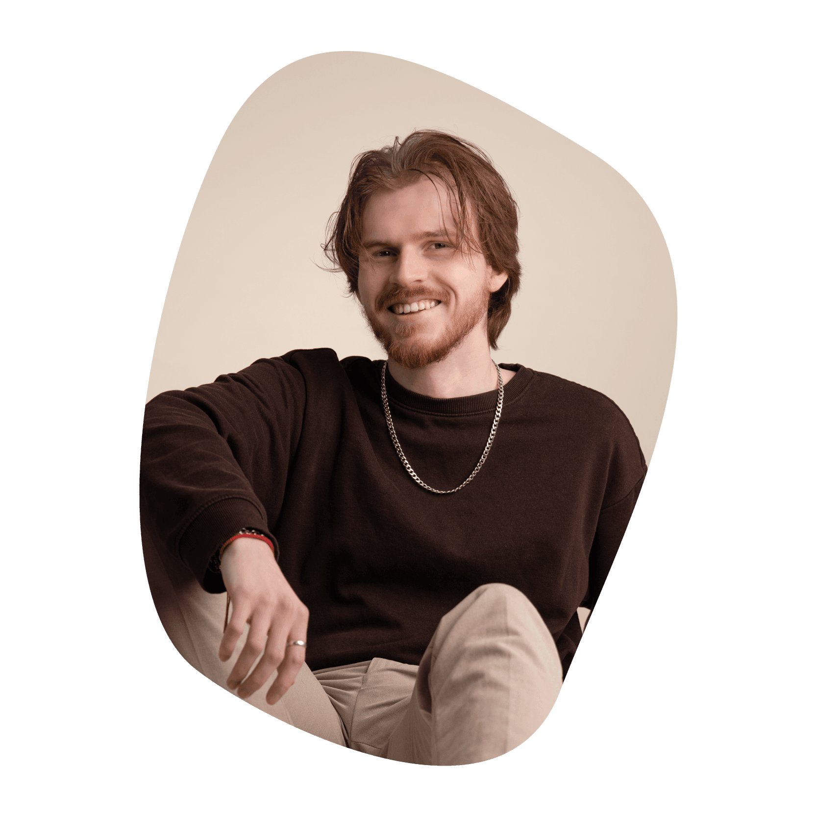 Un homme souriant aux cheveux mi-longs brun clair et à la barbe, portant un pull sombre, un pantalon clair et un collier à chaîne, est assis, détendu, sur un fond clair.