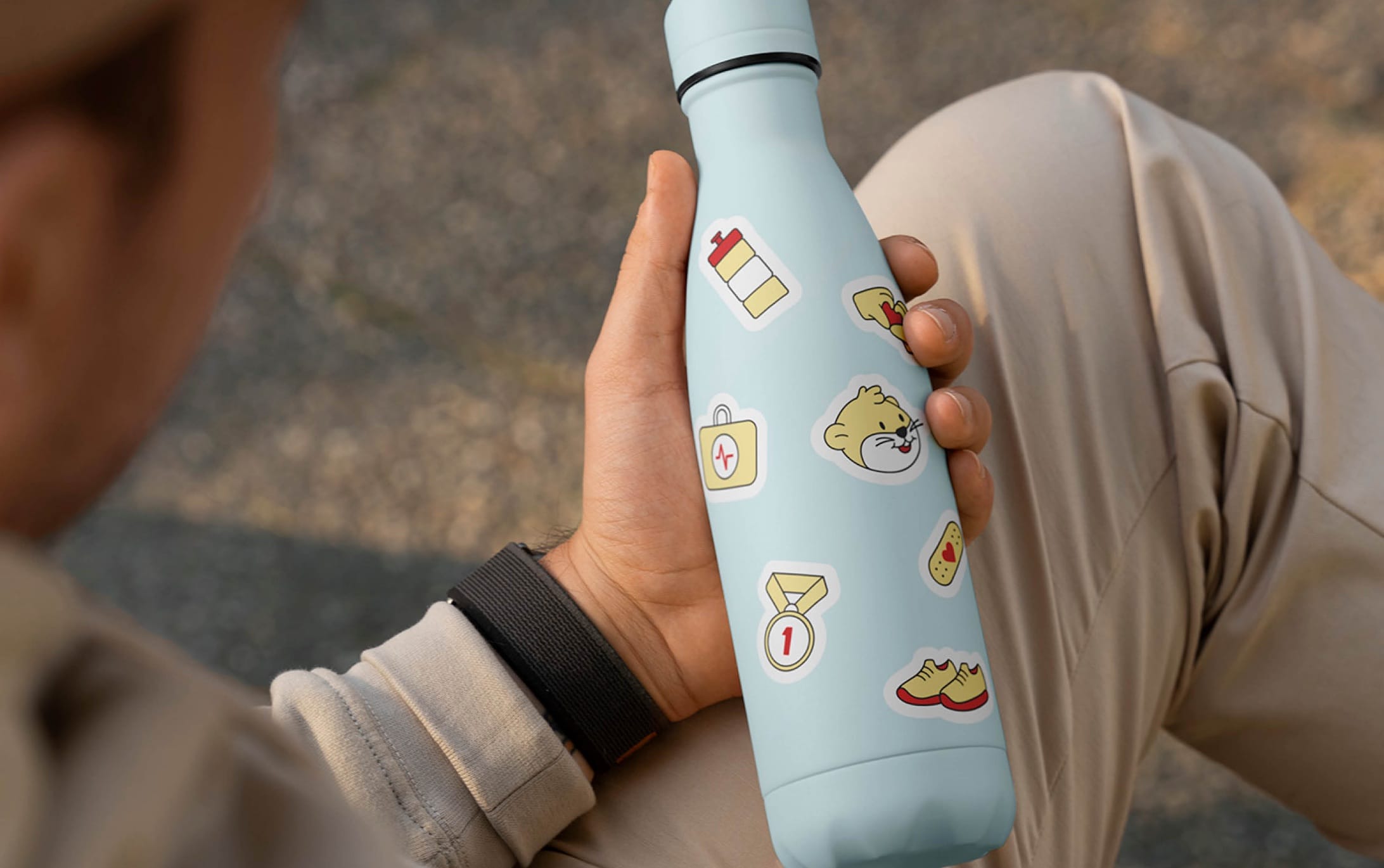 Gourde isotherme bleu clair couverte de stickers Charity Kids Run, tenue en main par un participant assis au bord d'un chemin.