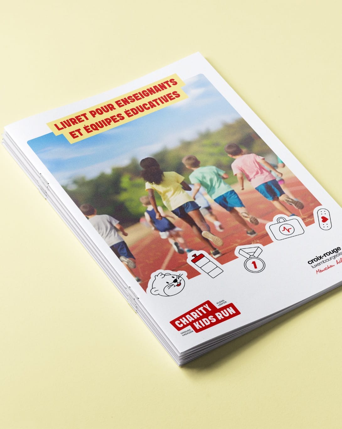 Couverture du livret pour enseignants et équipes éducatives : des enfants courent sur une piste, encadrés par les pictogrammes Charity Kids Run, sur fond jaune beurre.