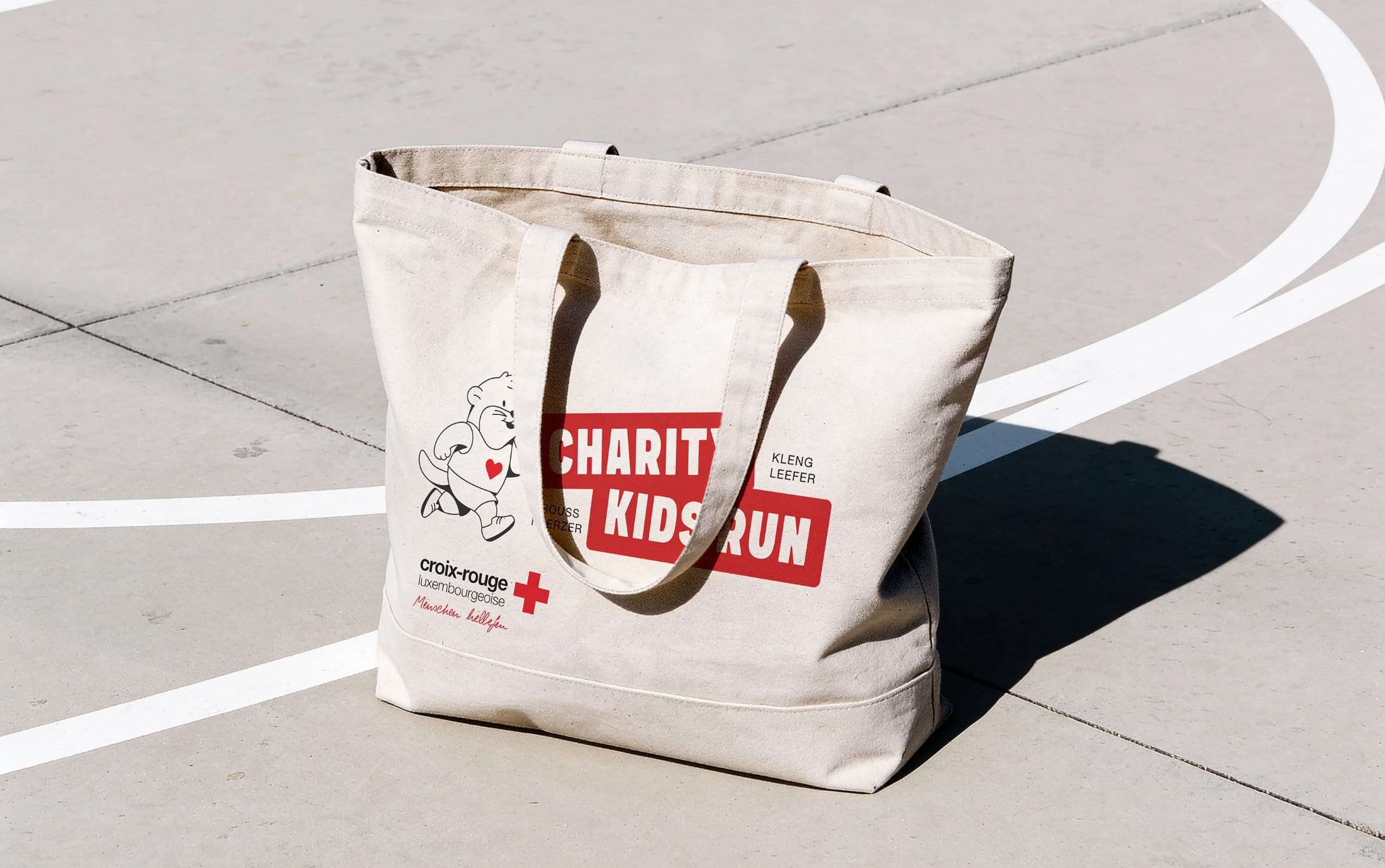 Tote bag en coton naturel siglé Charity Kids Run et Croix-Rouge luxembourgeoise, posé sur un terrain de sport en béton marqué de lignes blanches.