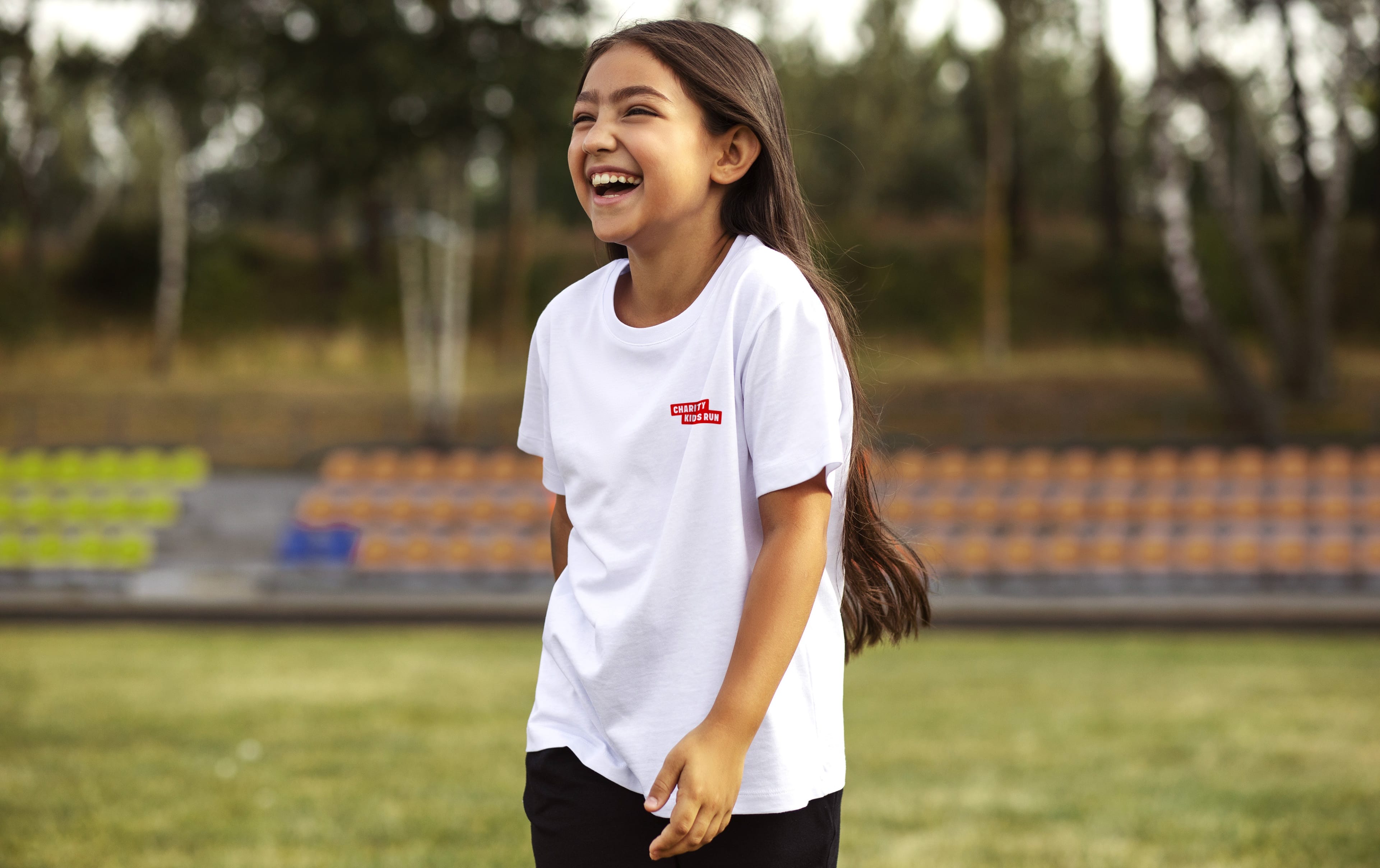Fillette riant aux éclats devant les gradins d'un stade, vêtue du t-shirt blanc floqué du logo Charity Kids Run.